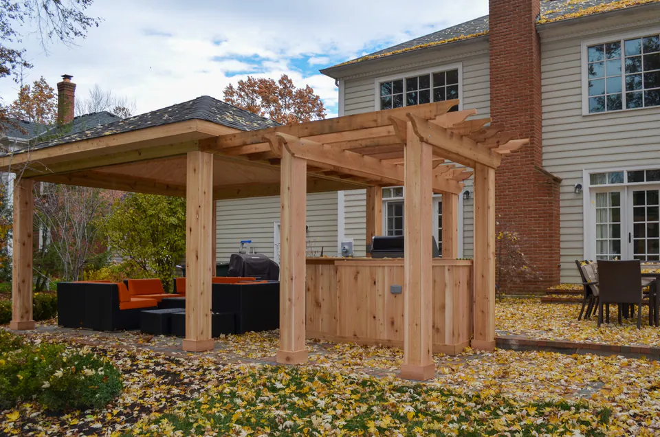 Exterior view of wood pavilion and outdoor kitchen beside house with paver patio, grill, lounge seating, and fall yard.